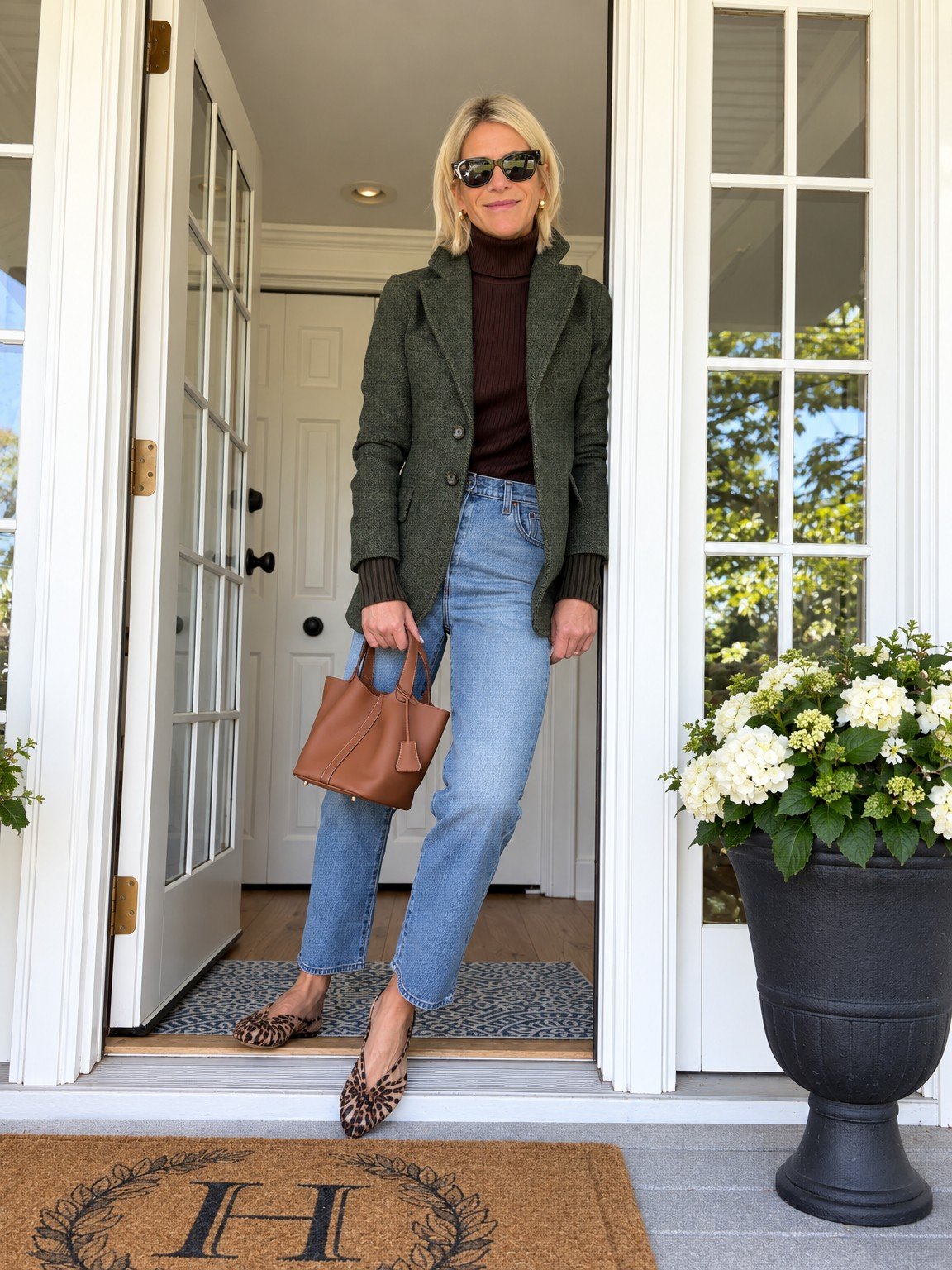 Angela in a green jacket and jeans with leopard slingbacks, holding a brown bag in a doorway.