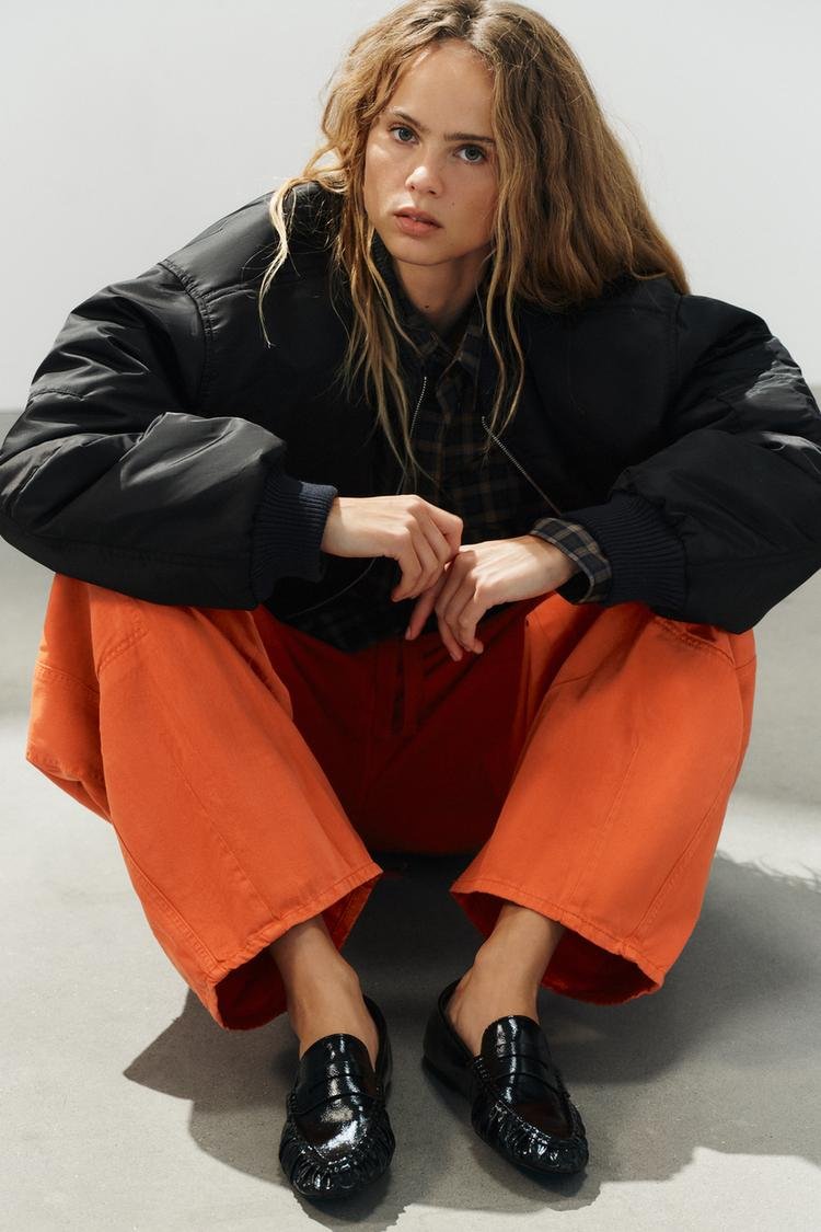 Zara model wearing black patent-effect gathered loafers with orange trousers.
