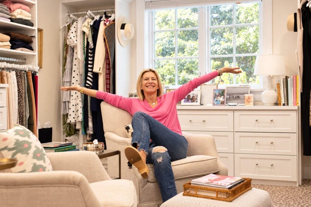 Angela Howenstein wearing a pink sweater and jeans with her arms outstretched in her closet.