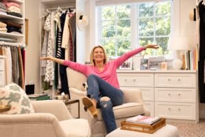 Angela Howenstein wearing a pink sweater and jeans with her arms outstretched in her closet.
