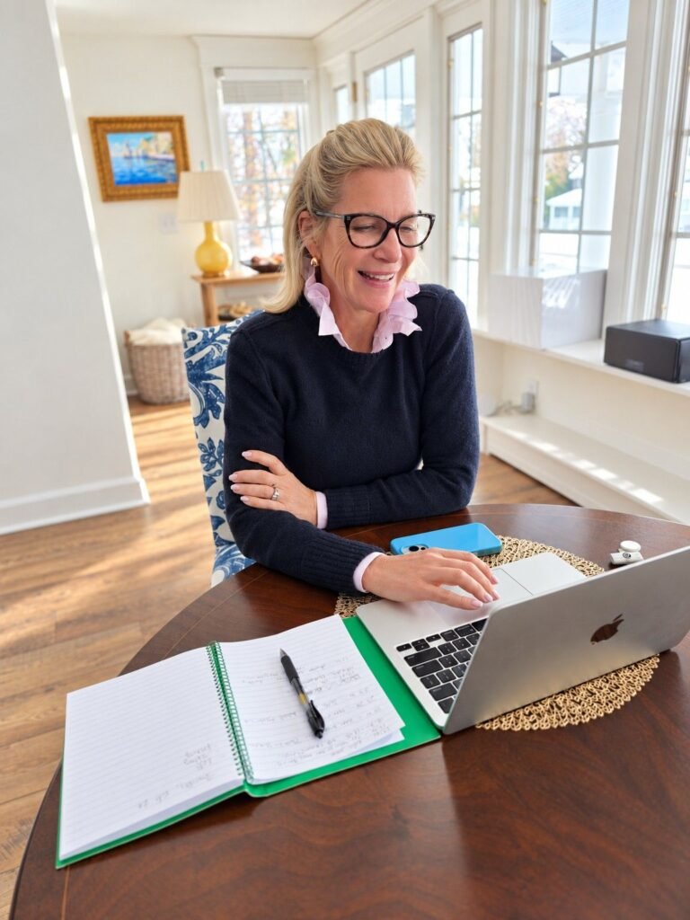 Angela Howenstein wearing a navy sweater over a white collared shirt while working at a laptop with an open notebook.