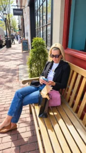 Angela Howenstein wearing a black blazer over a striped top with jeans and tan flats while seated on a bench.