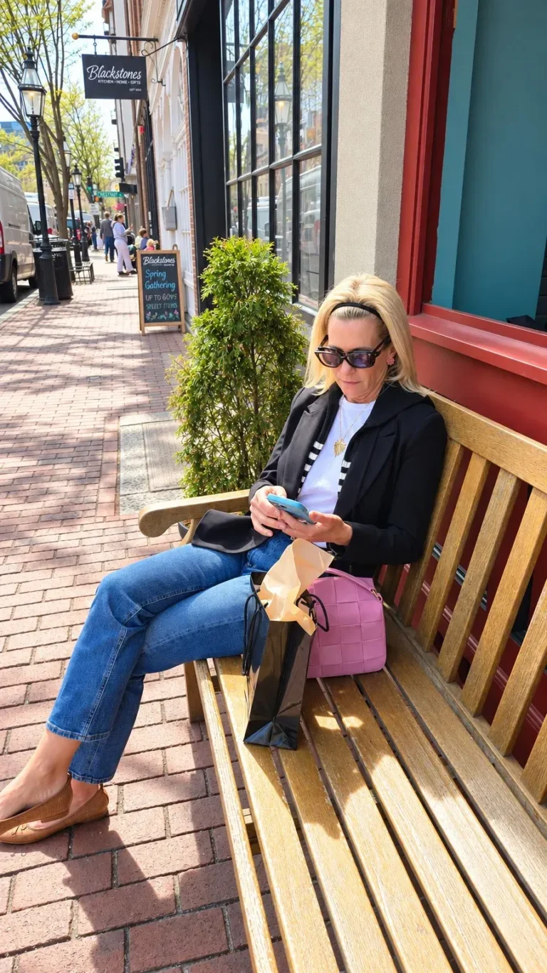 Angela Howenstein wearing a black blazer over a striped top with jeans and tan flats while seated on a bench.