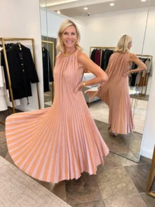 Angela Howenstein wearing a peach pleated halter dress and statement necklace in a fitting room.