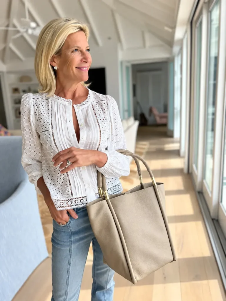 Angela Howenstein wearing a white eyelet blouse with jeans and carrying a taupe tote beside a window.
