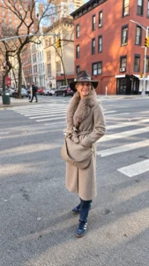 Angela Howenstein wearing a long taupe coat, dark hat and boots on a city street.