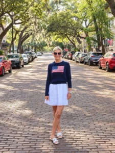 Angela Howenstein wearing a navy flag sweater, white skirt and flat sandals on a tree-lined street.