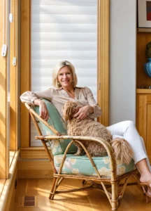 Angela Howenstein wearing a neutral sweater and white pants while seated with her dog in a wicker chair.