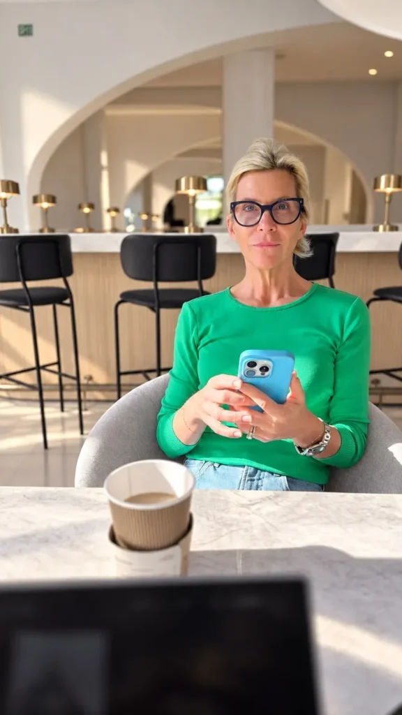 Angela Howenstein wearing a bright green sweater and glasses while seated at a cafe table.