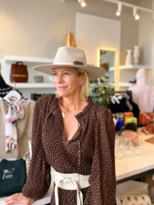 Angela Howenstein wearing a brown printed blouse, cream belt and wide-brimmed hat in a clothing boutique.