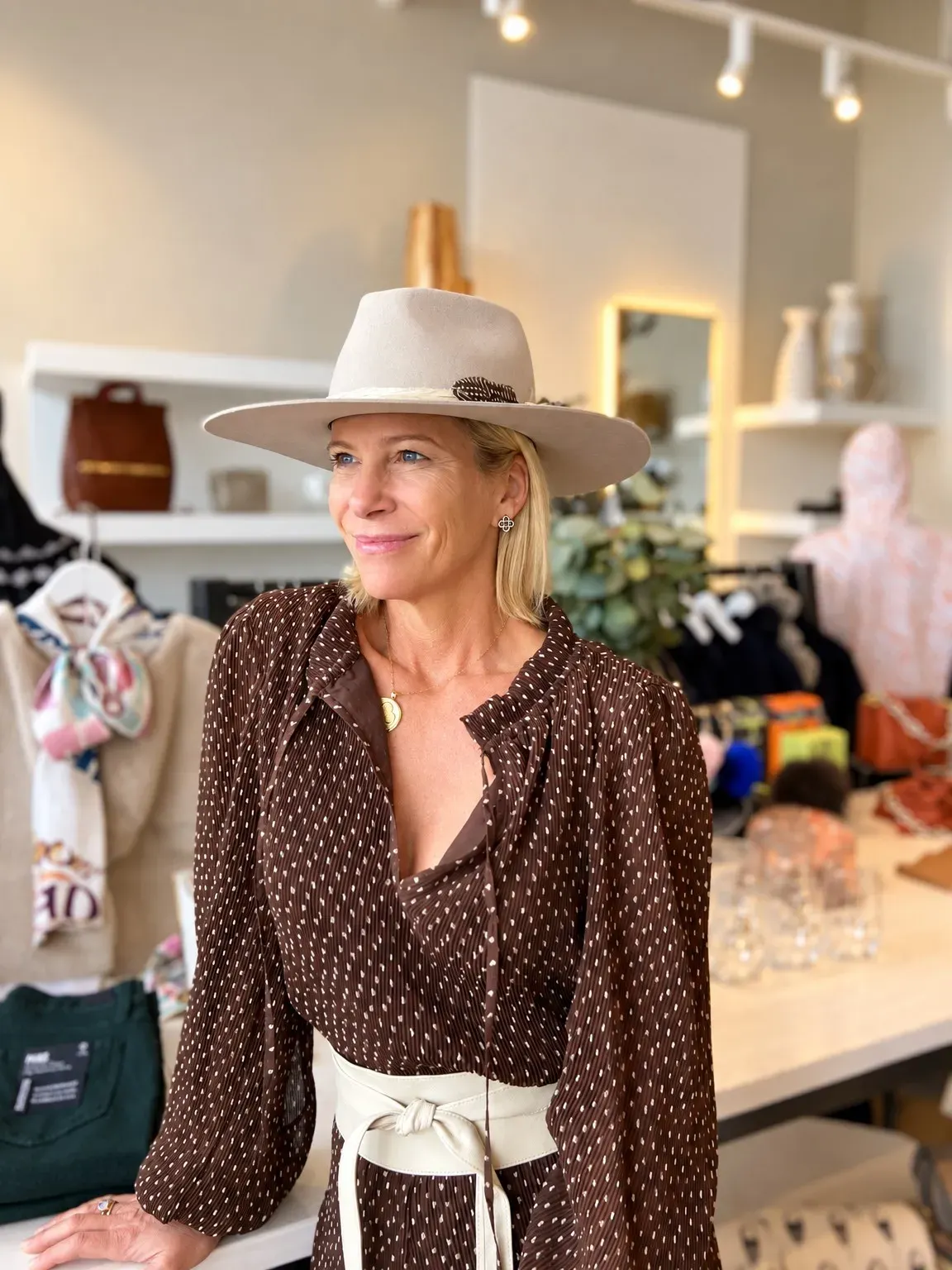 Angela Howenstein wearing a brown printed blouse, cream belt and wide-brimmed hat in a clothing boutique.
