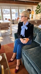 Angela Howenstein wearing a dark velvet blazer, striped tie-neck blouse, cuffed jeans and burgundy heels.