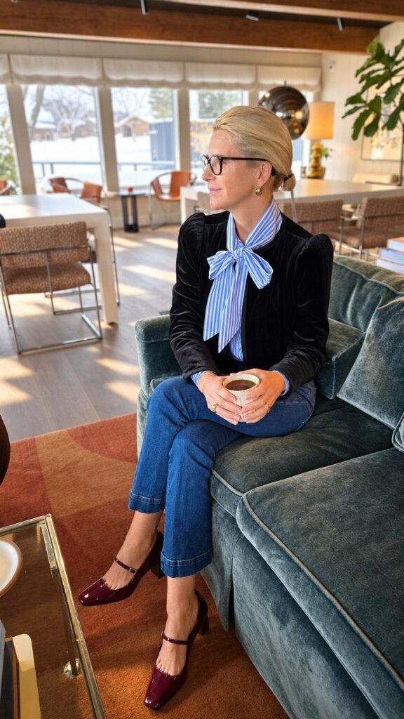Angela Howenstein wearing a dark velvet blazer, striped tie-neck blouse, cuffed jeans and burgundy heels.
